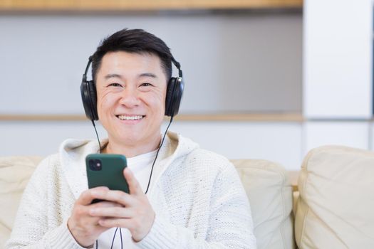 close up portrait of young asian man listening to music online in headphones using cellphone sitting on sofa at home