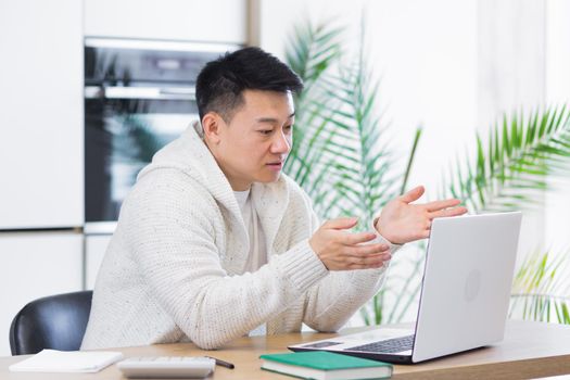 young asian man sitting at home in the living room or kitchen and communicating online. Male waving hand at laptop webcam. Video call learning distance. Training, conference or meeting E-learning