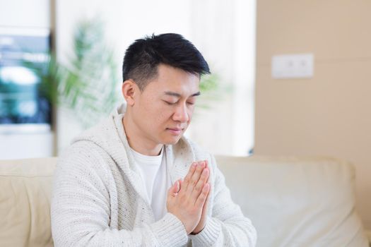 young asian man praying at home alone. Male folding pencil arms and meditating in apartment room. Christian religion traditional spiritual for their hope and believed god forgievness at house