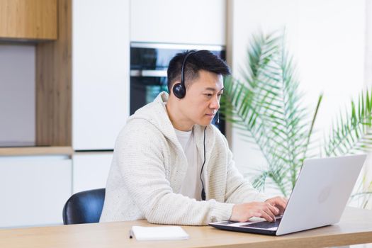 young asian man freelancer sitting at home at office desk with headset and talking online on video link via laptop in room or kitchen. The male indoors in casual clothes does the work and talks