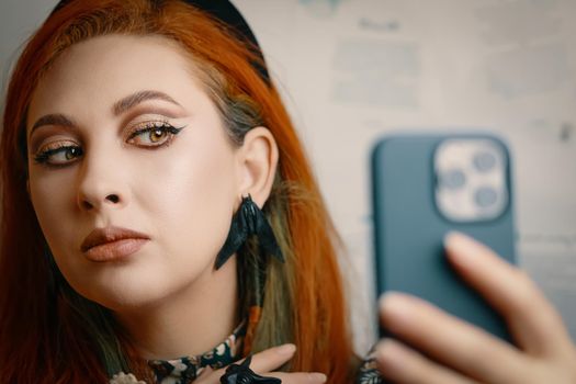 Female blogger runs a blog about fashion and makeup. Red-haired girl takes selfie on front camera of smartphone. Woman with Gothic-style jewelry with proud expression on her face. Witch aesthetic.