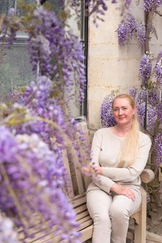 a beautiful middle-aged woman sits on a bench in the thickets of blooming wisteria and meditates happy. High quality photo