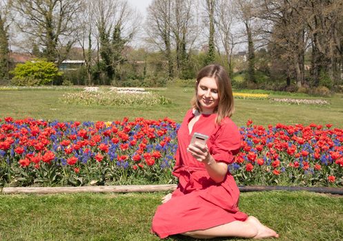 A young beautiful girl in a red dress in a spring park plays with her hair, flirts and takes a selfie on a mobile phone, tulips bloom and people walk in the park, pamper themselves.High quality photo