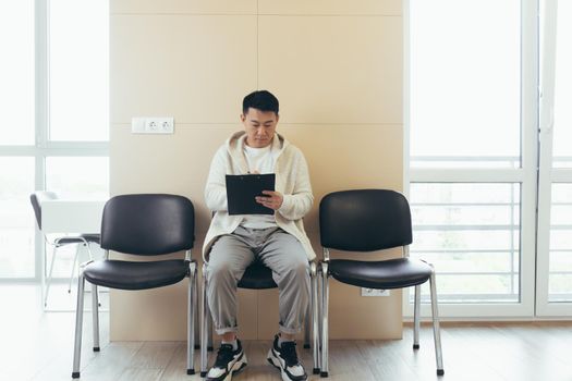 young asian male in waiting room for interview or meeting holding paper while sitting at chair fills out a resume questionnaire in office