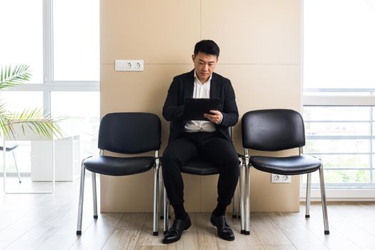 Asian businessman man, in the waiting room sitting on a chair near the reception of the office center, and fills out a test form to receive his application