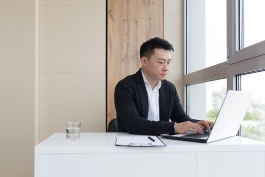 young asian office worker suffering from headache at work in the office drinks medicine pill with water. Sick man in a suit at the computer indoors with severe pain ache uses the drug in workplace