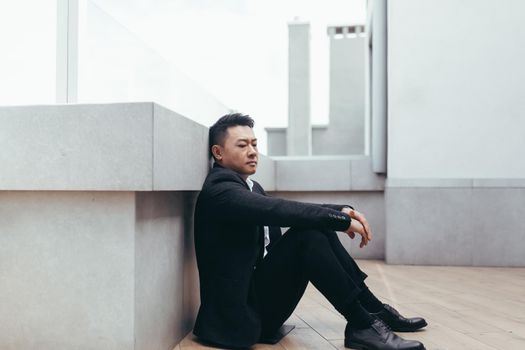 young asian depressed man sitting on outdoors street near office business center building. The upset male businessman lost his job due to the financial crisis and quarantine. Businessman has problems