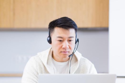 young asian man freelancer sitting at home at office desk with headset and talking online on video link via laptop in room or kitchen. The male indoors in casual clothes does the work and talks