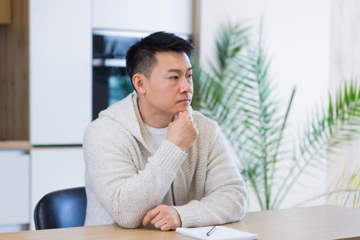 Thoughtful serious young asian man looking away. student writer sit at home office desk with laptop thinking of inspiration search solution ideas lost in thoughts dreaming solving financial problems