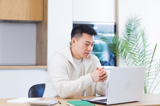 Young asian man working at home on a laptop computer sitting at a table in the kitchen