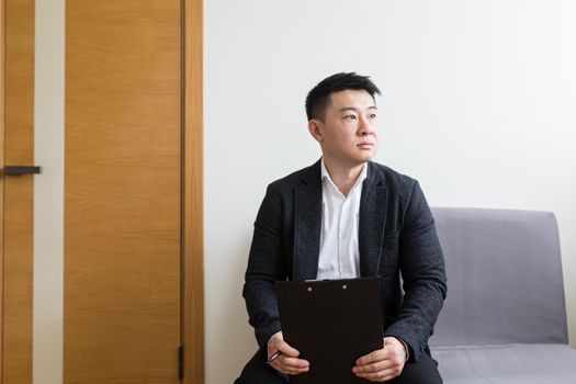 Young successful Asian man, waiting for a job interview, in the waiting room of the office center, sitting on a chair near the reception of the office center