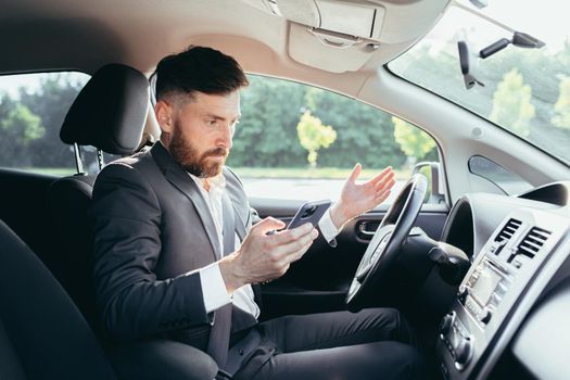 Male businessman sitting behind the wheel of a car, frustrated can not understand the steering and instructions funky car, reads additional information from the phone