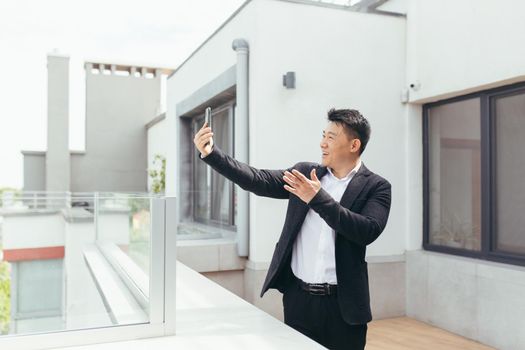 Successful asian businessman man talking on video phone, standing on balcony terrace of modern office center, happy and smiling