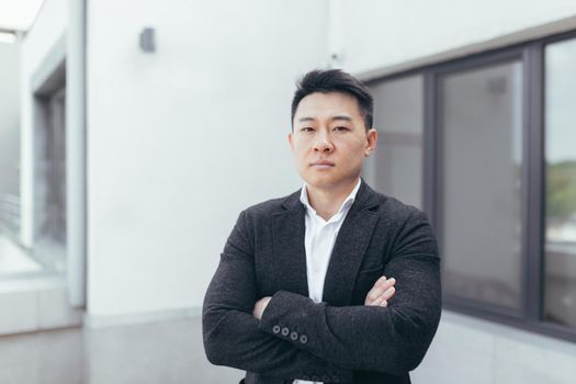 Young Asian businessman looks at the camera in a business suit, arms folded crosswise near the office center