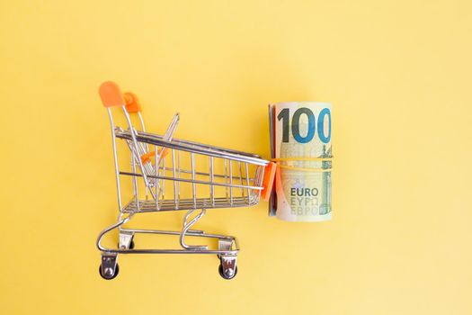 A roll of euro banknotes with a yellow elastic band in a small trolley on a yellow background. cash paper currency, payment, earning and savings, european currency money and finance concept. copy text