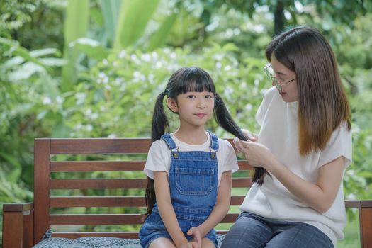 Asian smiling child schoolgirl with long dark hair and her mom in the morning, Happy family beautiful mother woman brushing her daughter hair while sitting outdoor at home, hairstyle care concept