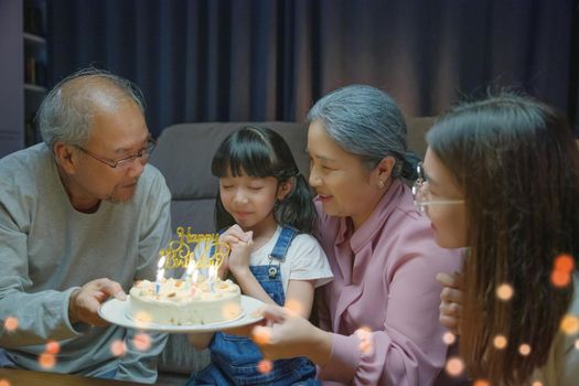 Happy Asian family grandparents and mother celebrating enjoy singing birthday song party with cake of granddaughter blowing candle at home in night time, Birthday surprise concept