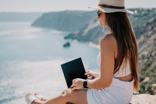 Successful business woman in yellow hat working on laptop by the sea. Pretty lady typing on computer at summer day outdoors. Freelance, travel and holidays concept.