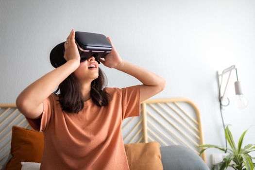 Teen girl using VR headset at home. Amazed Asian young female wearing virtual reality goggles. Modern technology and lifestyle concepts.