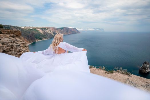 Blonde with long hair on a sunny seashore in a white flowing dress, rear view, silk fabric waving in the wind. Against the backdrop of the blue sky and mountains on the seashore