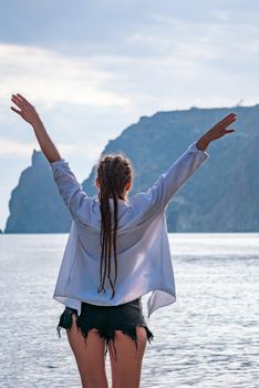 The girl stands on the shore and looks at the sea. Her hands are raised up. She wears a white shirt and her hair is in a braid