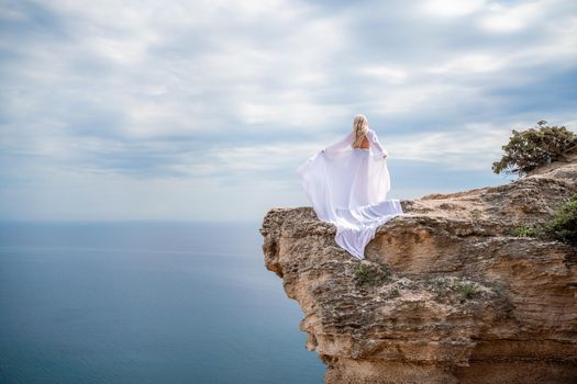 Blonde with long hair on a sunny seashore in a white flowing dress, rear view, silk fabric waving in the wind. Against the backdrop of the blue sky and mountains on the seashore