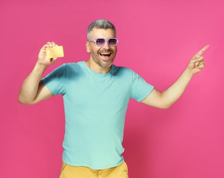 Great offer, handsome man hold debit, credit card in hand pointing sideways wearing blue t-shirt and glasses isolated on pink background. Man with bank card in hand. Financial, banking concept.