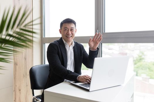 Successful Asian businessman, in a black business suit, works on a laptop, in a stylish office explains a new business strategy via video link