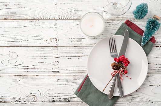 Christmas table setting with white dishware, silverware and red and green decorations on white wooden background. Top view, flat lay copy space