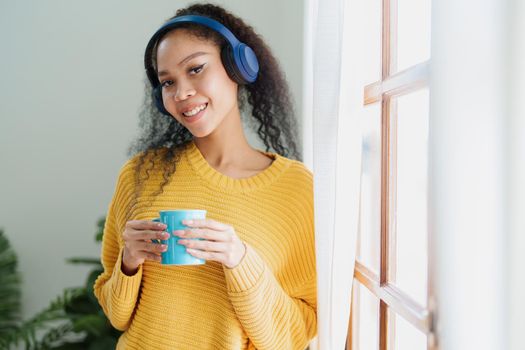 African American wearing headphones and holding coffee mugs smiling happily while listening to music relaxing at home