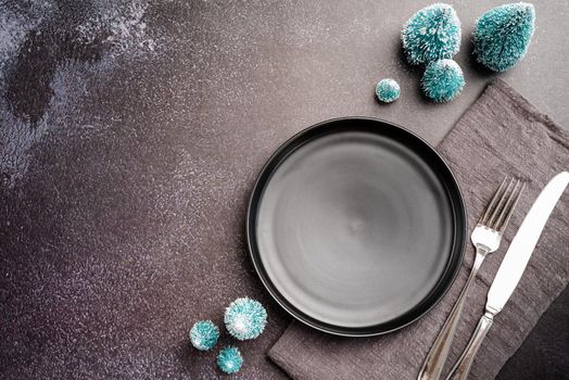 Christmas table setting with black dishware, silverware and green decorations on dark background. Top view flat lay
