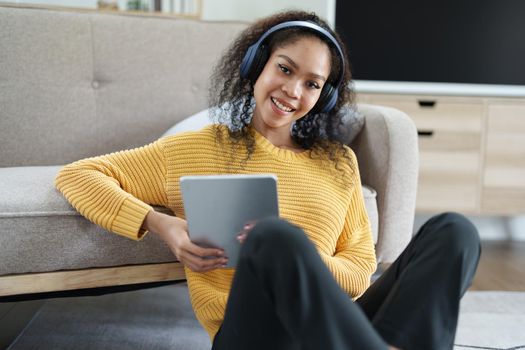 Portrait of an African American with headphones and tablet smiling happily listen music while relaxing on the sofa at home.