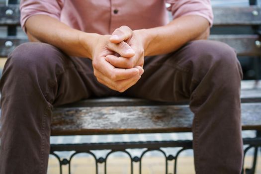 No face visible. Man spend time sitting on the bench with hands put together wearing pink shirt. Overpassing problems young man sits outdoors thinking on life moments. Psychology concept.
