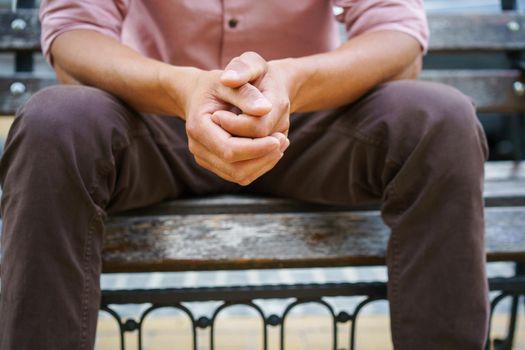 No face visible. Man spend time sitting on the bench with hands put together wearing pink shirt. Overpassing problems young man sits outdoors thinking on life moments. Psychology concept.