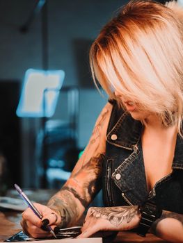 Tattoo artist drawing illustration of skull inside ink studio. Woman with black nails and rings at work. New fashion lifestyle artistic trends concept . Warm cinematic filter.