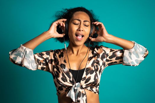 Beautiful african american woman with afro hair and big headphones having fun smiling and dancing in studio against blue background. High quality photo