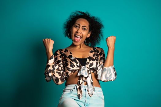 Surprised excited happy african woman on blue background. Girl with curly hair and positive trendy appearance shows yes gesture of victory,she achieved result, goals. High quality photo