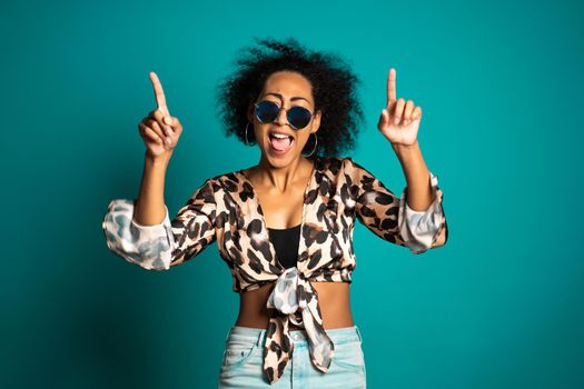 Pretty african woman with afro hair in leopard top and sunglasses smiling to camera over blue wall background. Cute mixed race girl's portrait. High quality photo