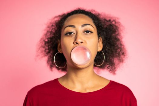 Playful woman in red wear blows bubblegum, chewing gum. Pretty african american lady stands on pink background. High quality photo