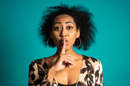 African american woman holding a finger on her lips over blue background. Gesture of shhh, secret, silence. High quality photo