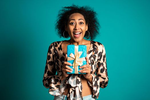 Excited african woman received gift box with bow. She is happy and flattered by attention. Girl smiling with present on blue background. Studio portrait. High quality photo