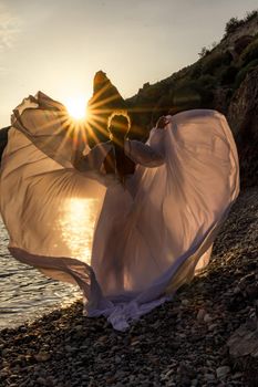 A mysterious female silhouette with long braids stands on the sea beach with mountain views, Sunset rays shine on a woman. Throws up a long white dress, a divine sunset