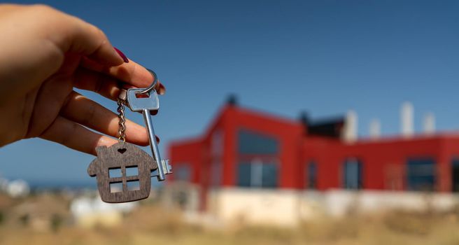 A woman's hand holds a house key against the backdrop of a house under construction. Real estate agent. Buying a house, apartment