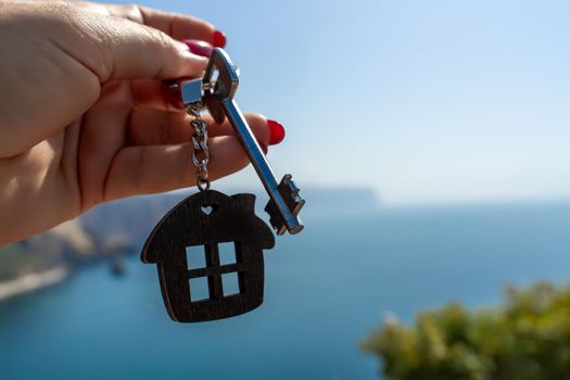 Keys with a keychain in the shape of a house in a female hand against the backdrop of the sea. The concept of buying a house, apartment, real estate, young family, mortgage loan, rental housing