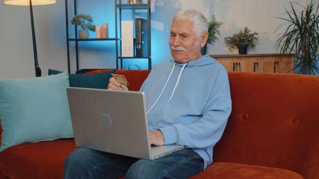 Portrait of old man using credit bank card and laptop computer while transferring money, purchases online shopping, order food delivery at home. Senior grandfather sitting on couch at home living room