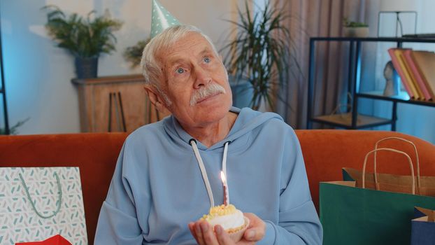 Happy senior grandfather wears festive birthday hat hold cupcake makes wish joyful congratulating blowing burning candle on cake. Mature man celebrating anniversary party on couch with gifts at home