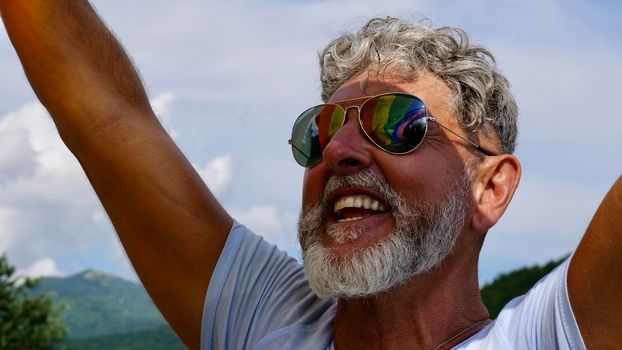 Portrait senior of a gray-haired elderly Caucasian man with a beard and sunglasses holding a rainbow LGBTQIA flag against a sky background in gay parade. Celebrates Pride Month, Coming Out Day