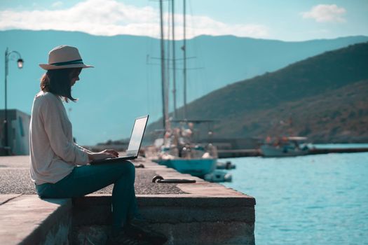 A young girl in a hat is sitting on the pier and working, typing on a laptop keyboard on a sunny day against a beautiful background of a seascape with moored yachts in the bay. Woman works and travels