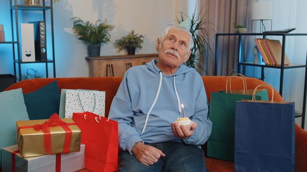 Happy senior grandfather wears festive birthday hat hold cupcake makes wish joyful congratulating blowing burning candle on cake. Elderly man celebrating anniversary party on couch with gifts at home