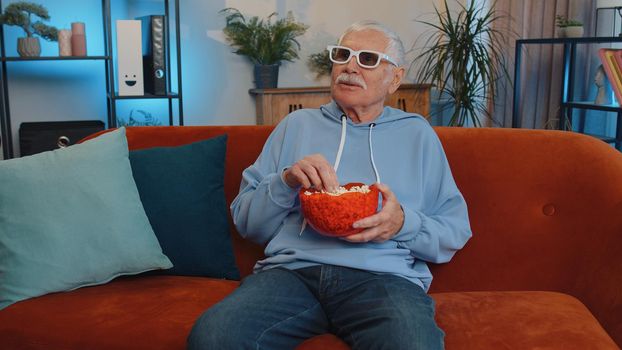 Excited elderly man on sofa eating popcorn and watching interesting tv serial, sport game, film, online social media movie content online at home. Senior grandfather enjoying domestic entertainment